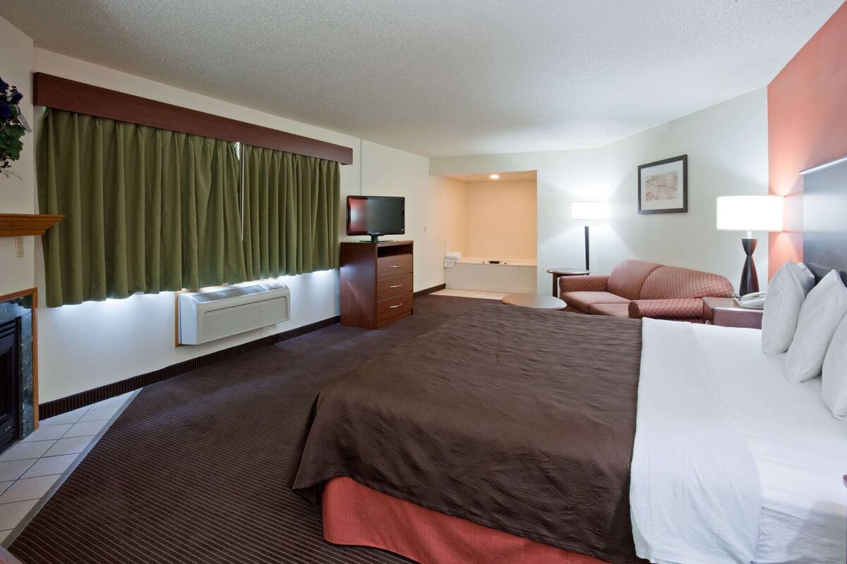 Habitación de hotel con una cama grande, un escritorio con computadora, un televisor y un ventanal con cortinas.