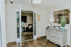 Ein Hotel-Rezeptionsbereich mit einem Schreibtisch, einem Sessel, einer Lampe, einem Willkommensschild und einem Fenster mit Jalousien.