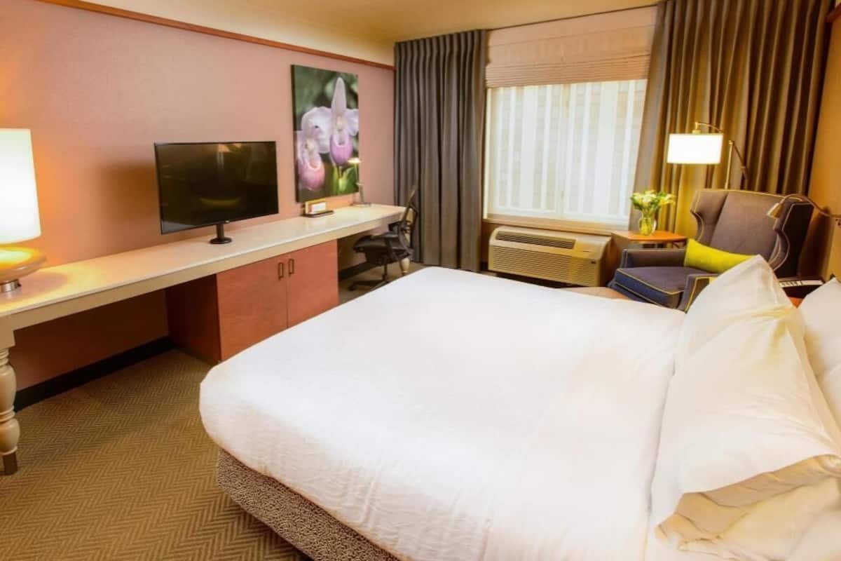 A hotel room with a bed, a desk with a television, a chair, a window with curtains, and an air conditioning unit.