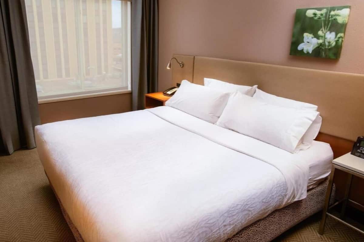 A hotel room with a large bed, a bedside table with a lamp, a small table with a clock, a window with curtains, and a wall-mounted picture.