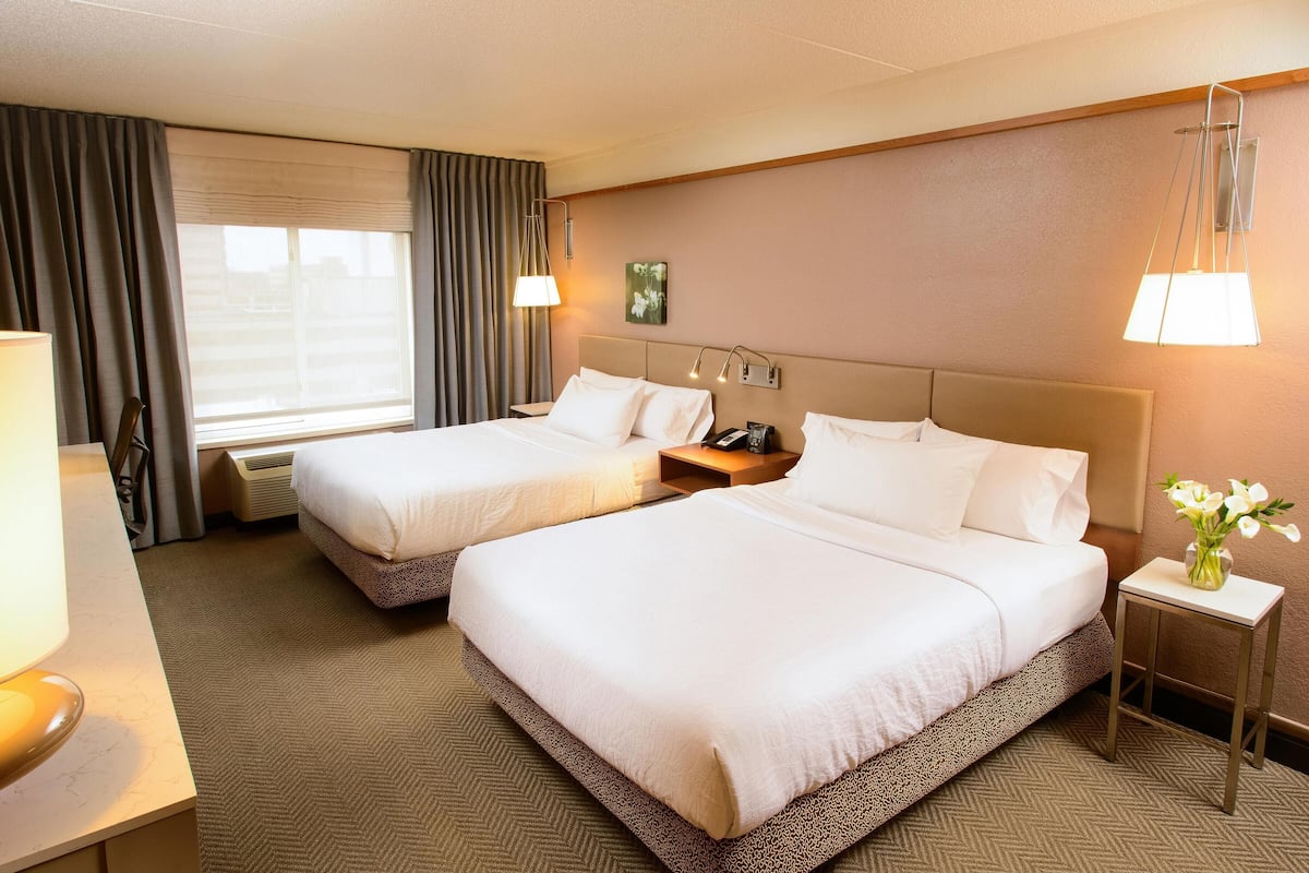 A hotel room with two beds, a desk, a lamp, a nightstand, and a vase with flowers.
