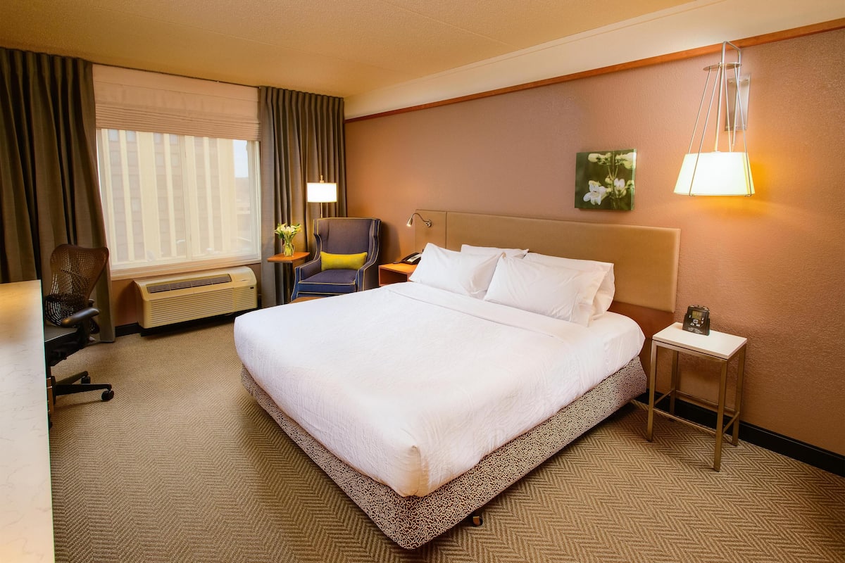 A hotel room with a large bed, a desk with a chair, a lamp, and a small table with a phone.