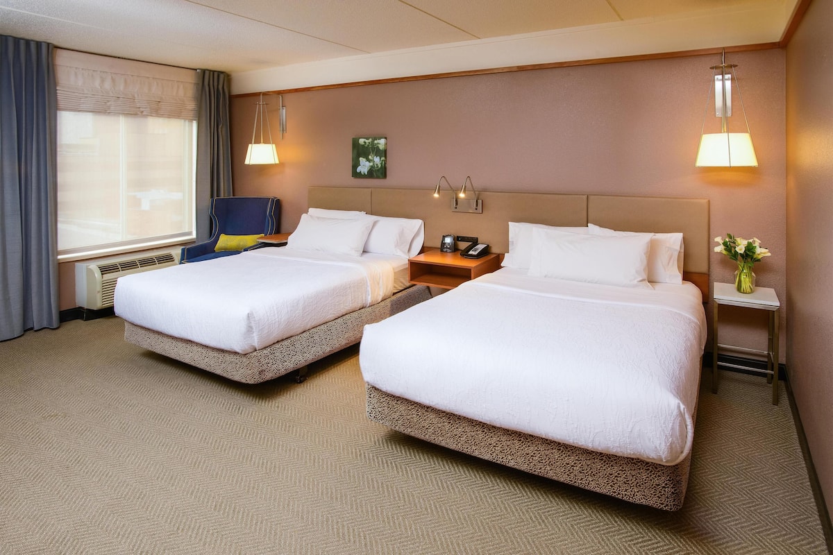 A hotel room with two beds, a small table, a chair, a nightstand, a lamp, and a window with curtains.