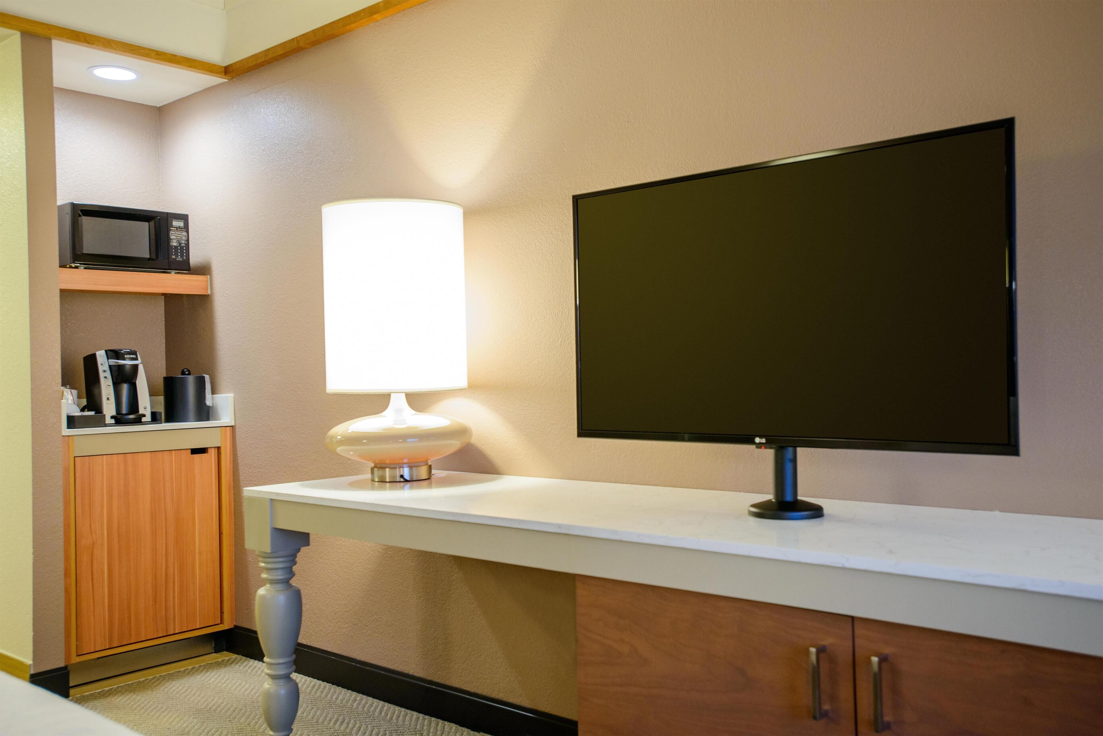 Une chambre d’hôtel dotée d’un téléviseur à écran plat fixé au mur, d’un four à micro-ondes placé au-dessus d’un meuble en bois et d’une table blanche avec une lampe.