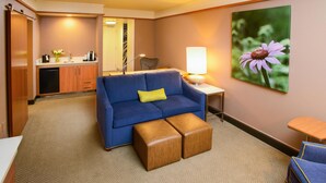 A hotel room with a blue sofa, a wooden bench, a small table with a lamp, a painting of a flower, and a kitchenette in the background.