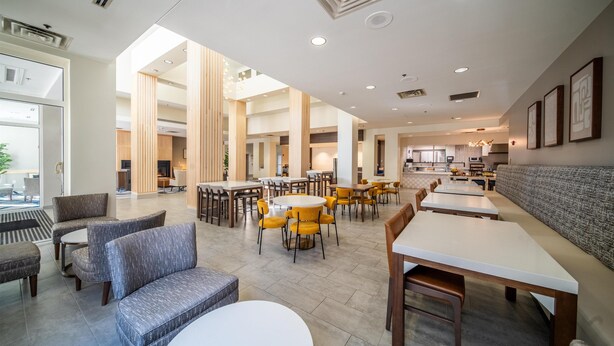 A spacious hotel lobby with multiple seating areas, including sofas and round tables, and a large dining area with yellow chairs.