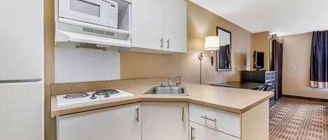 Cocina de hotel compacta con microondas, estufa, pileta y muebles de cocina blancos.