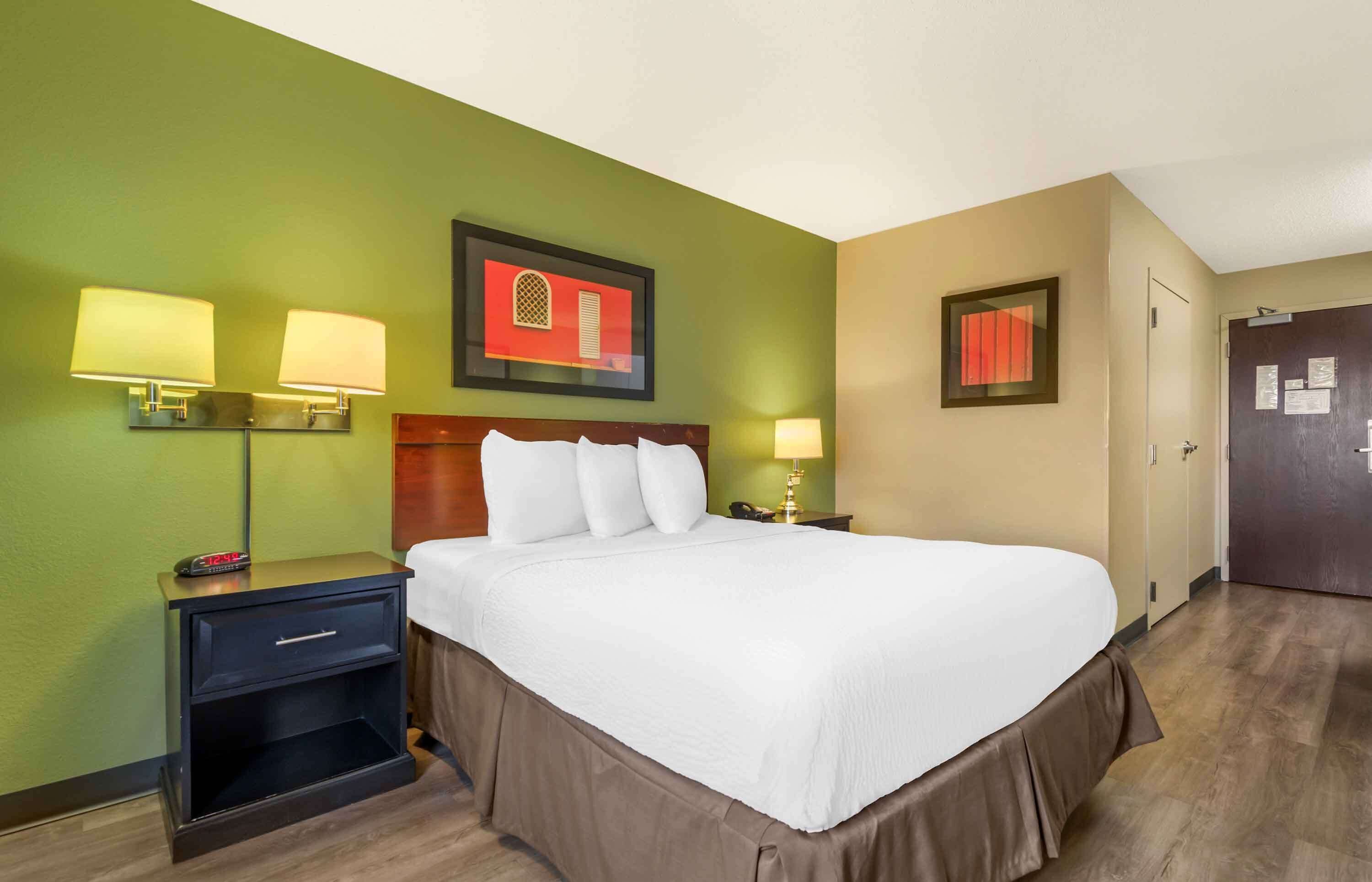 Une chambre d’hôtel avec un grand lit, deux tables de chevet avec des lampes, une tête de lit en bois et des murs de couleur verte et beige.