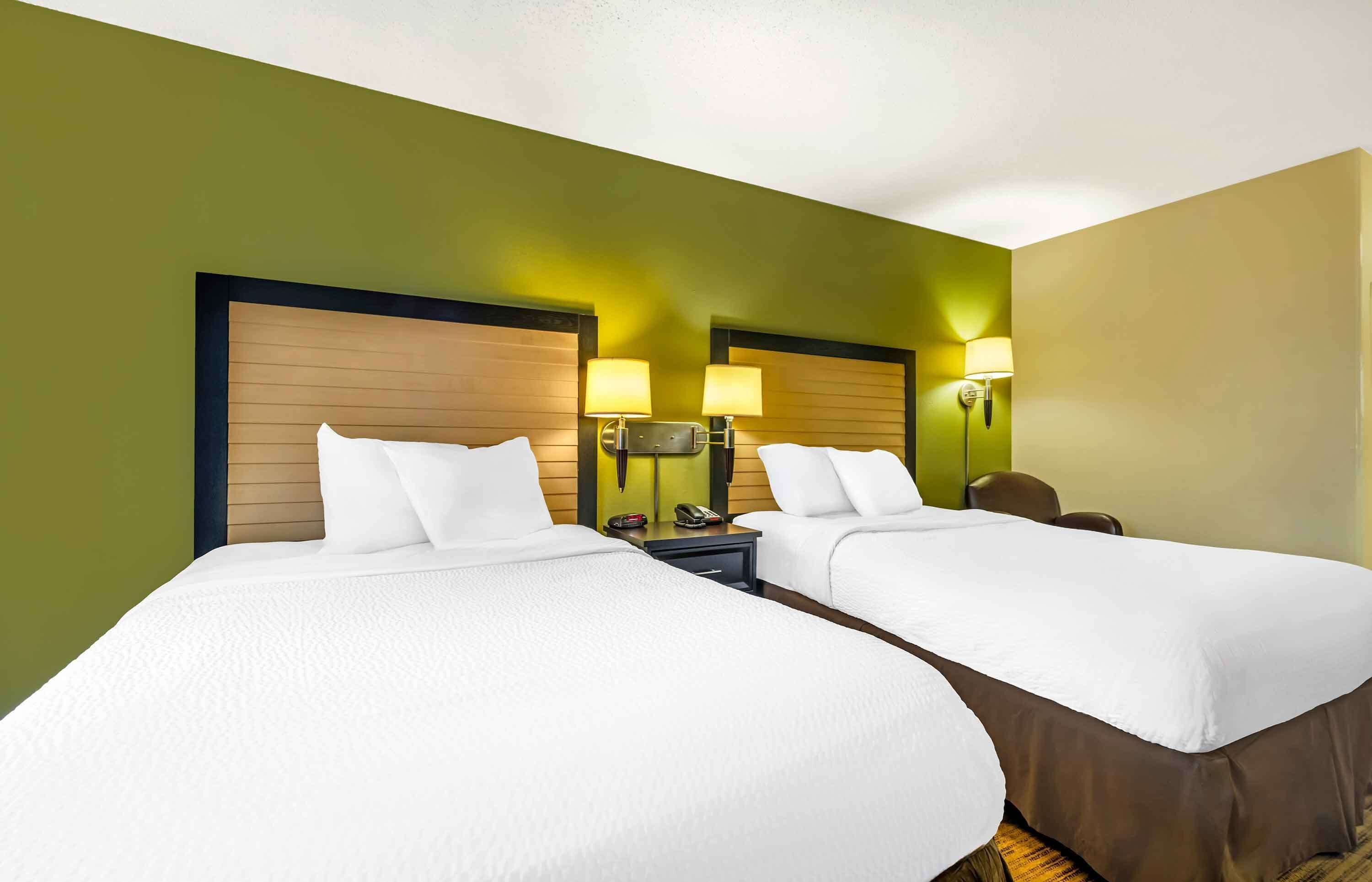 Habitación de hotel con dos camas, cada una con ropa blanca y cabeceros de madera. Hay lámparas de noche y un mesita de noche con un teléfono.