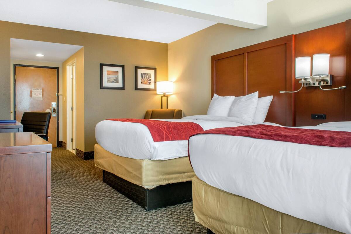 A hotel room with two beds, a desk, a chair, a lamp, and framed pictures on the wall.