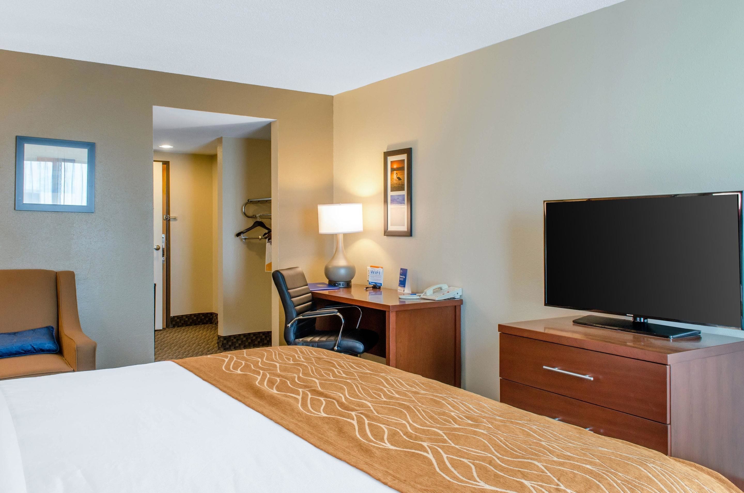 A hotel room with a bed, a desk with a lamp, a TV, and a chair.