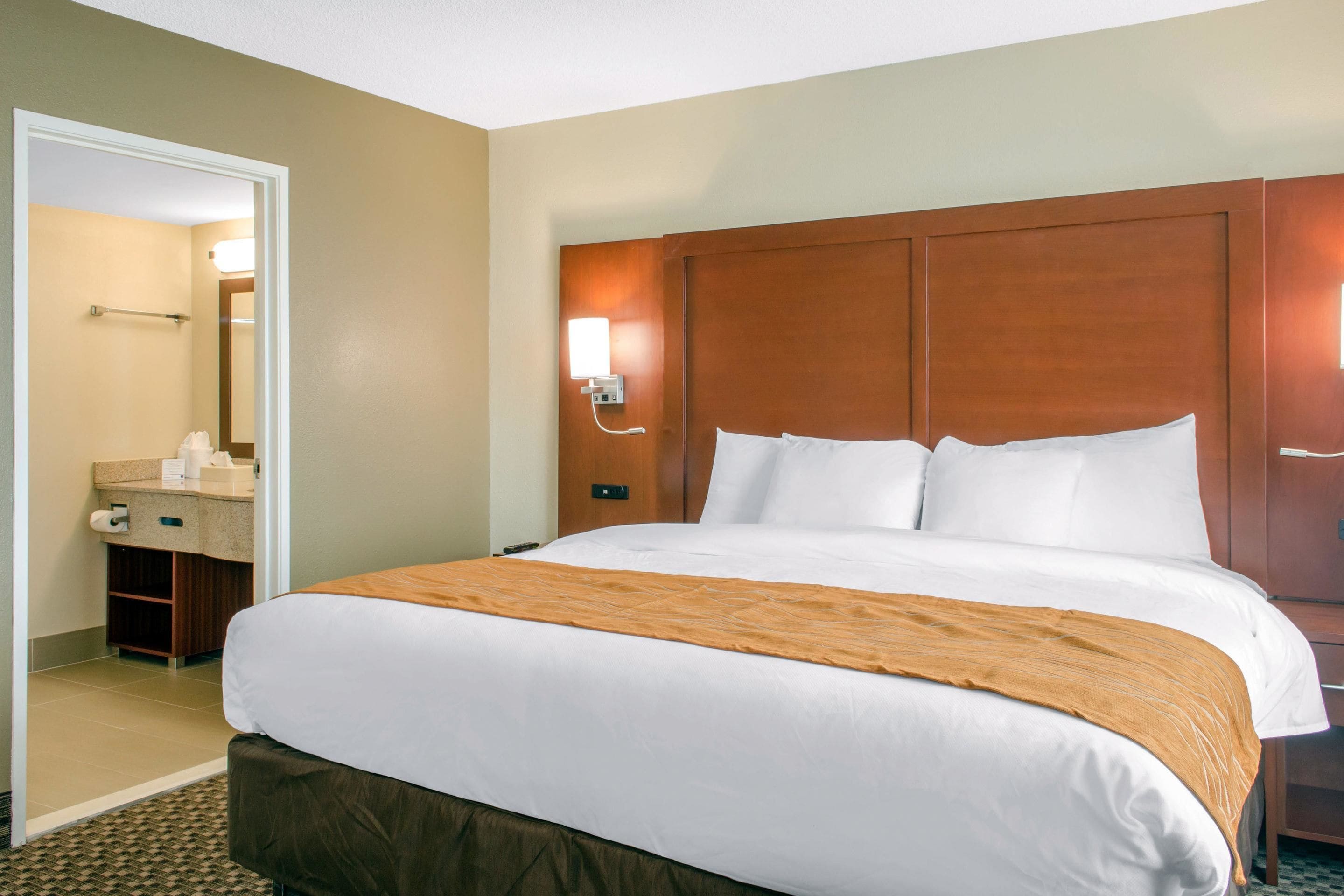 A hotel room with a large bed, a wooden headboard, a bedside table, a nightstand, a mirror, and a bathroom with a towel rack and toilet paper.