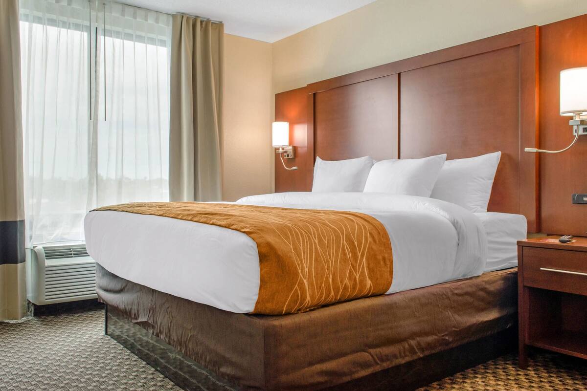 A hotel room with a large bed, a bedside table, a wall-mounted lamp, and a window with curtains.