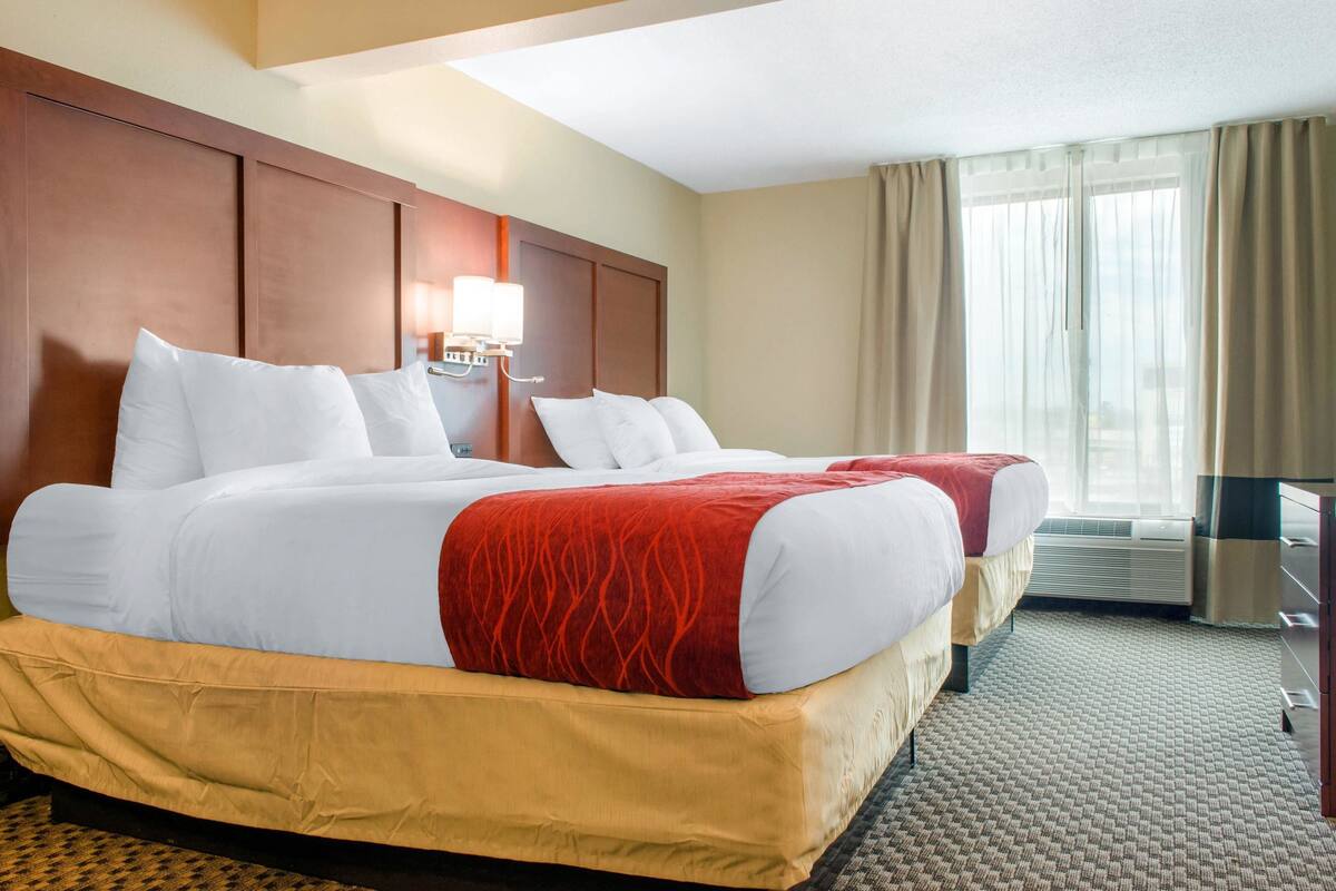 A hotel room with two beds, a wooden headboard, a wall-mounted lamp, and a large window with curtains.