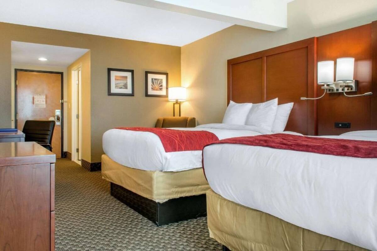 A hotel room with two beds, a desk, a chair, a lamp, and framed pictures on the wall.