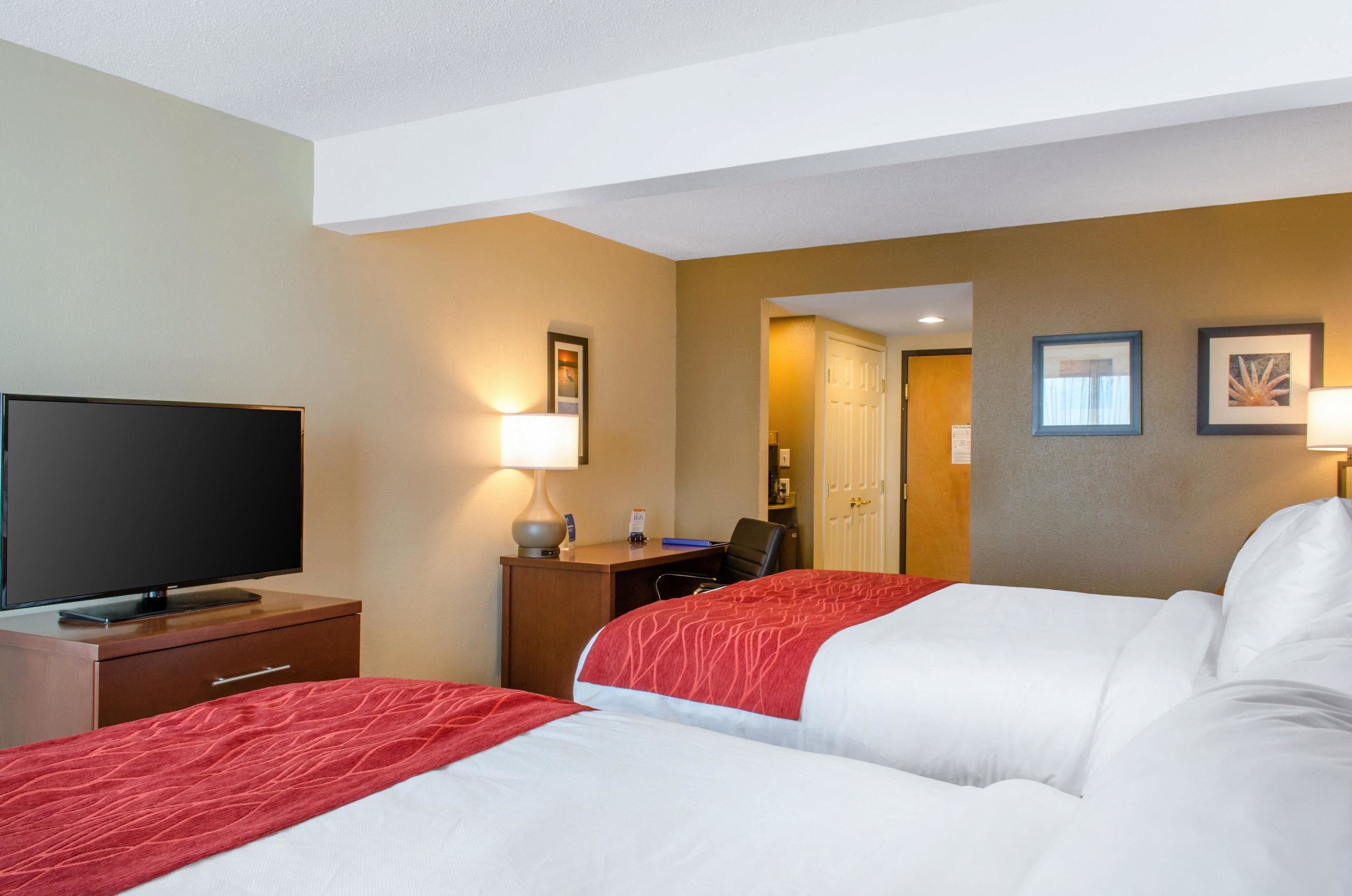 Une chambre d’hôtel avec deux lits, un bureau, une chaise, une télévision et une porte donnant sur une autre pièce.