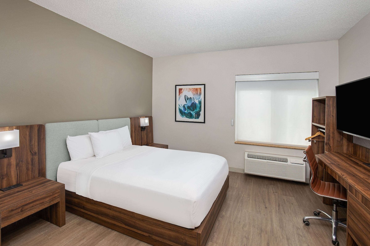 Una habitación de hotel con una cama grande, cabecera de madera, mesitas de noche con lámparas, un televisor de pantalla plana, un escritorio con silla y una ventana con persianas.