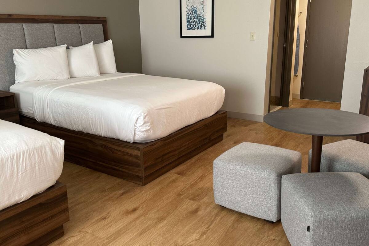 Un dormitorio moderno con una cama grande, una mesita pequeña y dos otomanas grises.