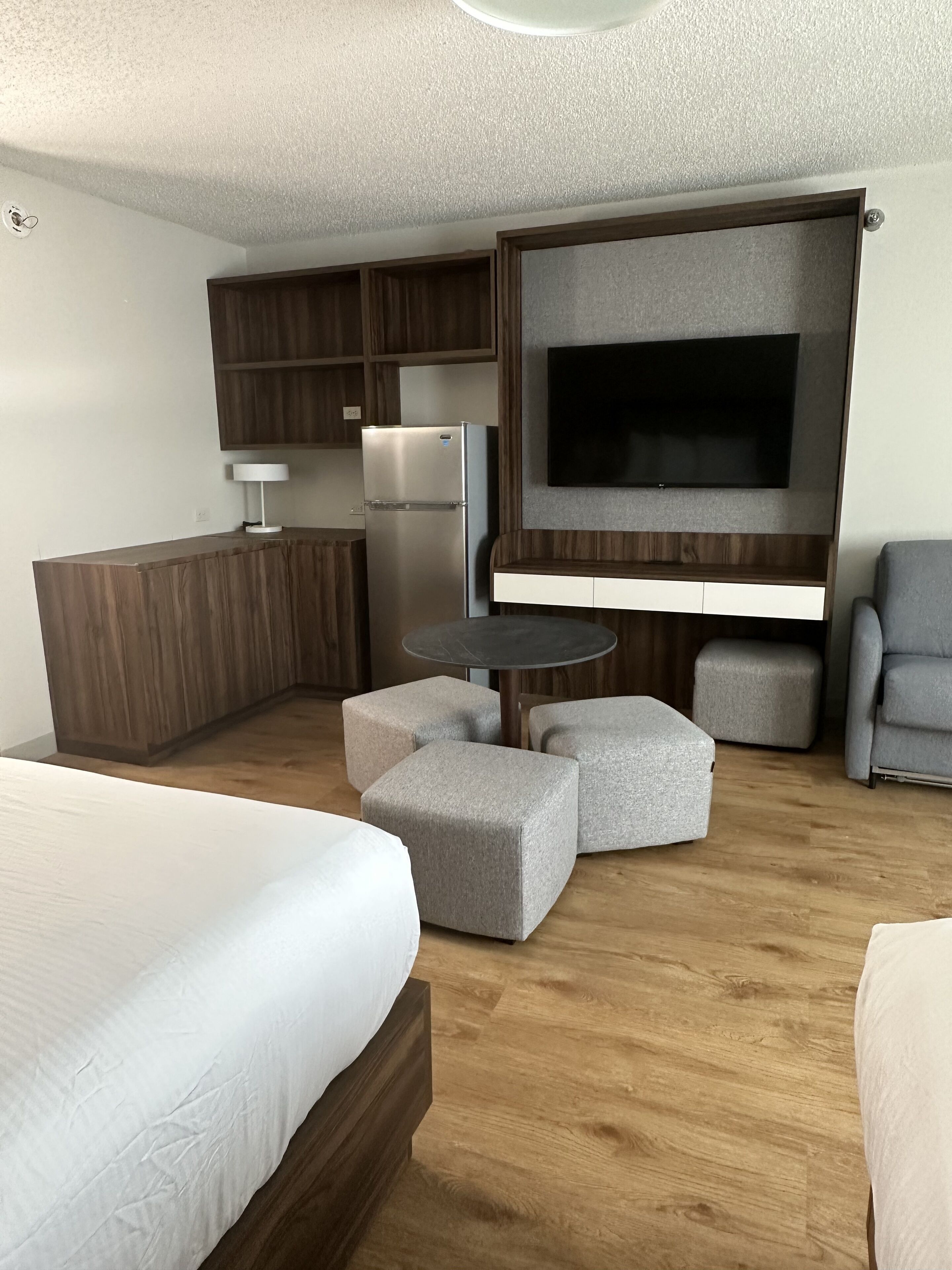 A modern hotel room with a bed, a kitchenette, a flat-screen TV, and a seating area with ottomans.