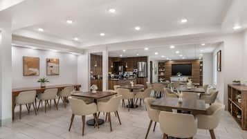 A spacious dining area with multiple tables and chairs, a kitchen in the background, and a large mirror reflecting the space.
