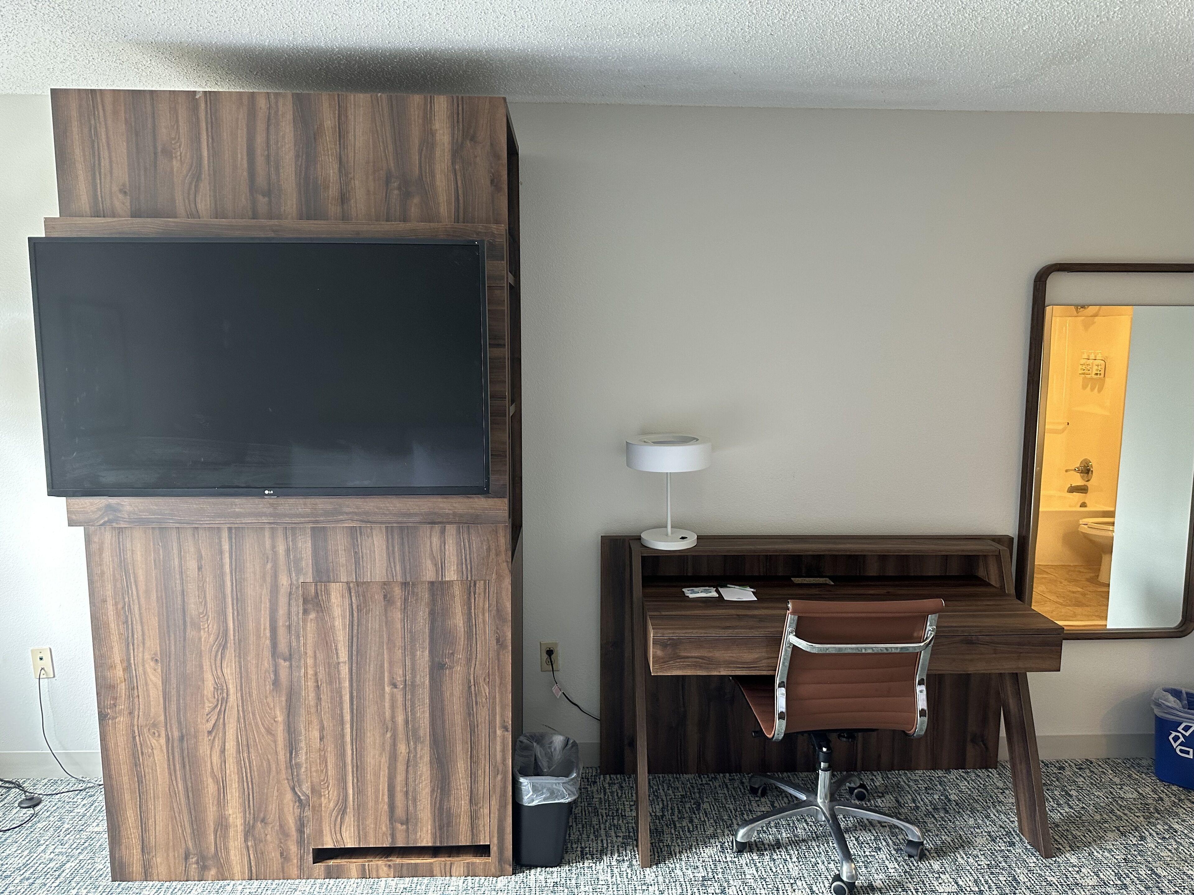 Habitación de hotel con un mueble bajo de madera para el televisor, un escritorio con una silla, una lámpara de escritorio blanca y un espejo grande en la pared.