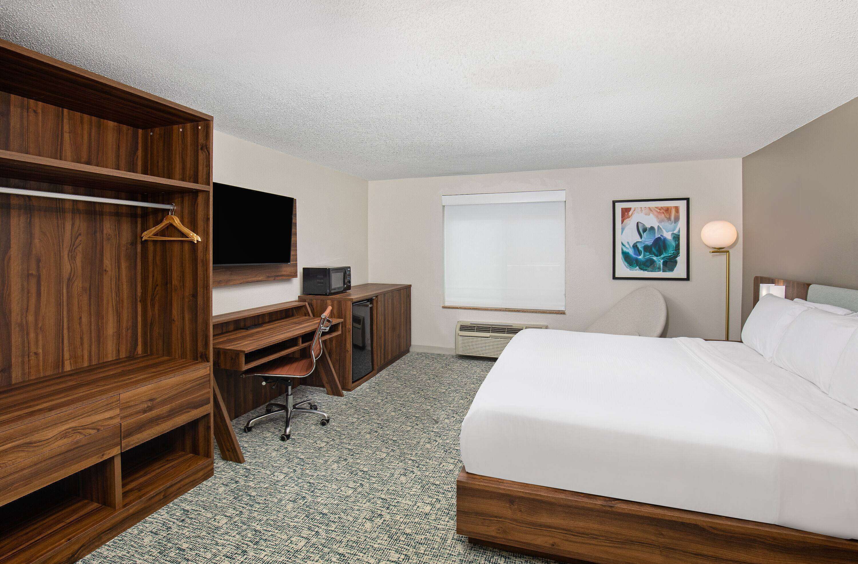 A hotel room with a wooden wardrobe, a desk with a chair, a bed, a flat-screen TV, a window with blinds, and a painting on the wall.