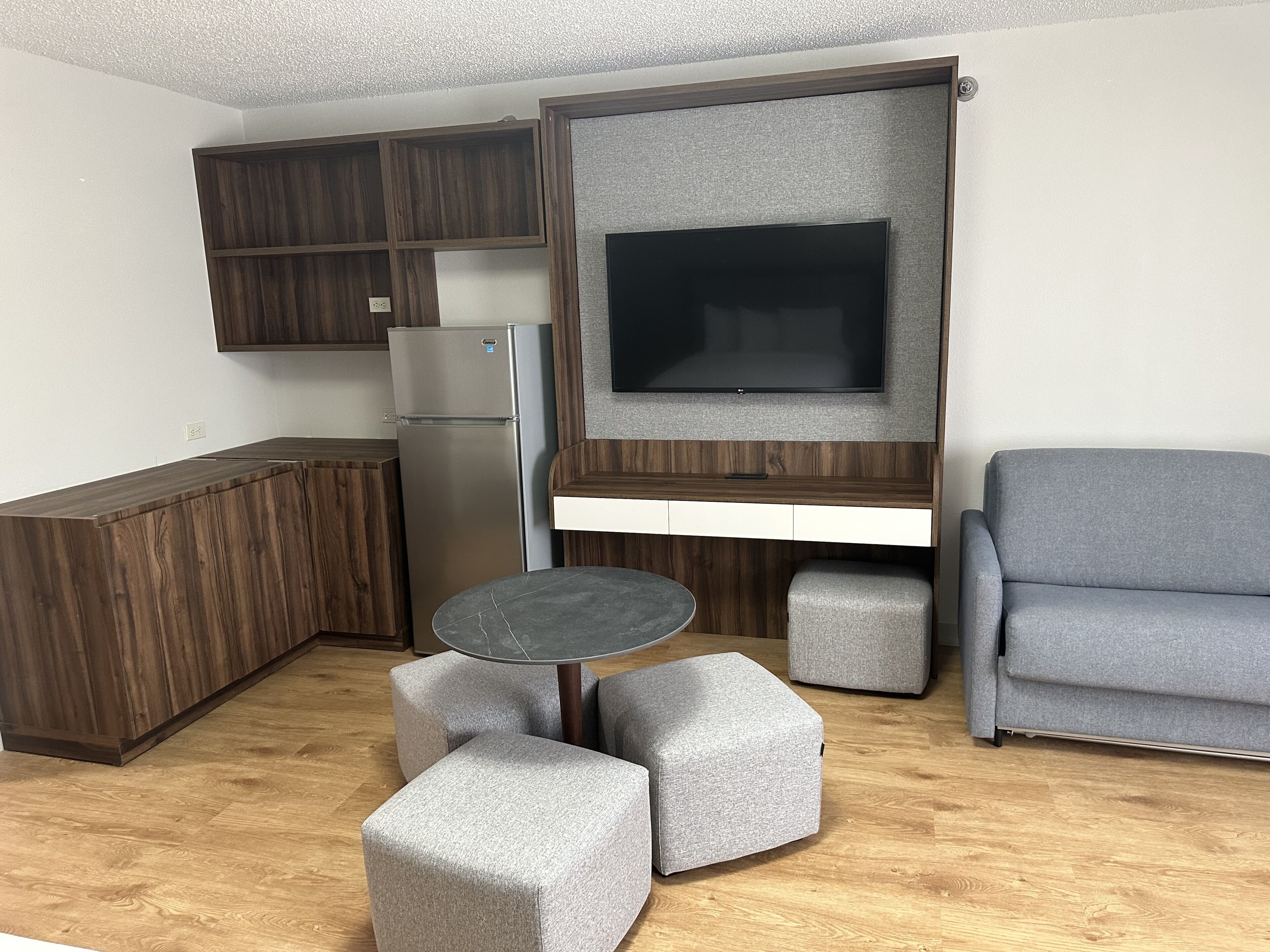 Un espace de vie compact comprenant un coin cuisine, un téléviseur à écran plat fixé sur un panneau en bois et un canapé gris avec des poufs.