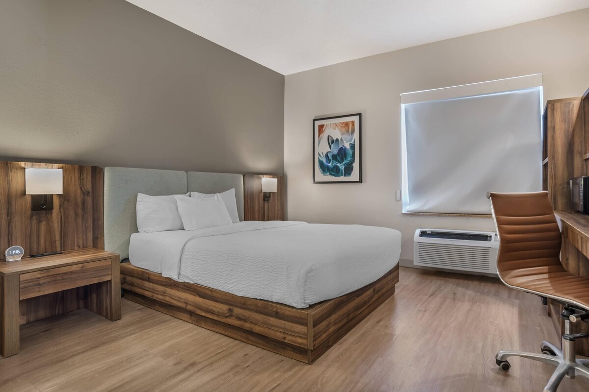 Una camera d'albergo con un letto, comodini, una scrivania, una sedia, una televisione, una finestra e un'opera d'arte appesa al muro.