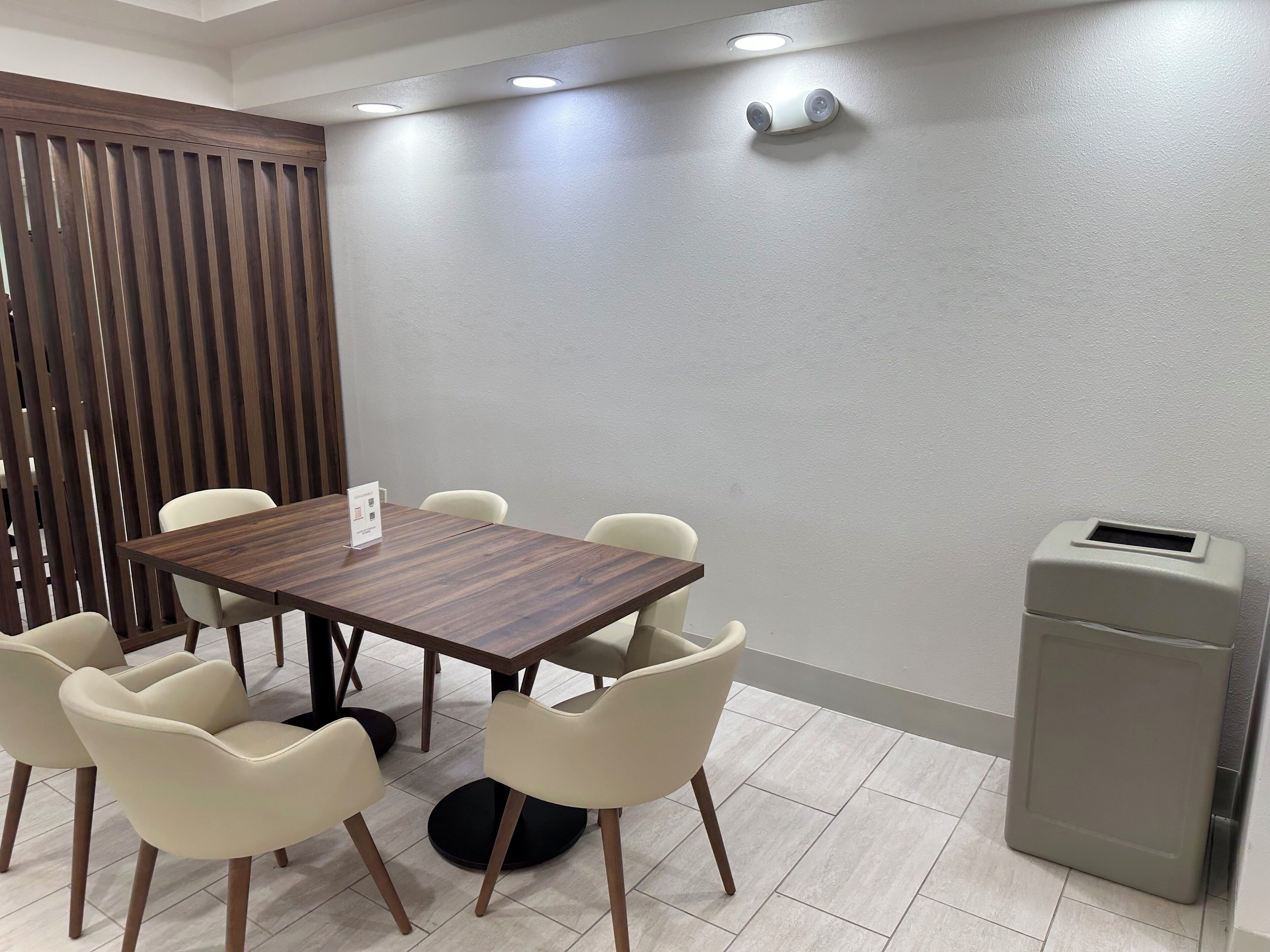 A conference room with a wooden table, six chairs, a small trash bin, and a wall-mounted light fixture.