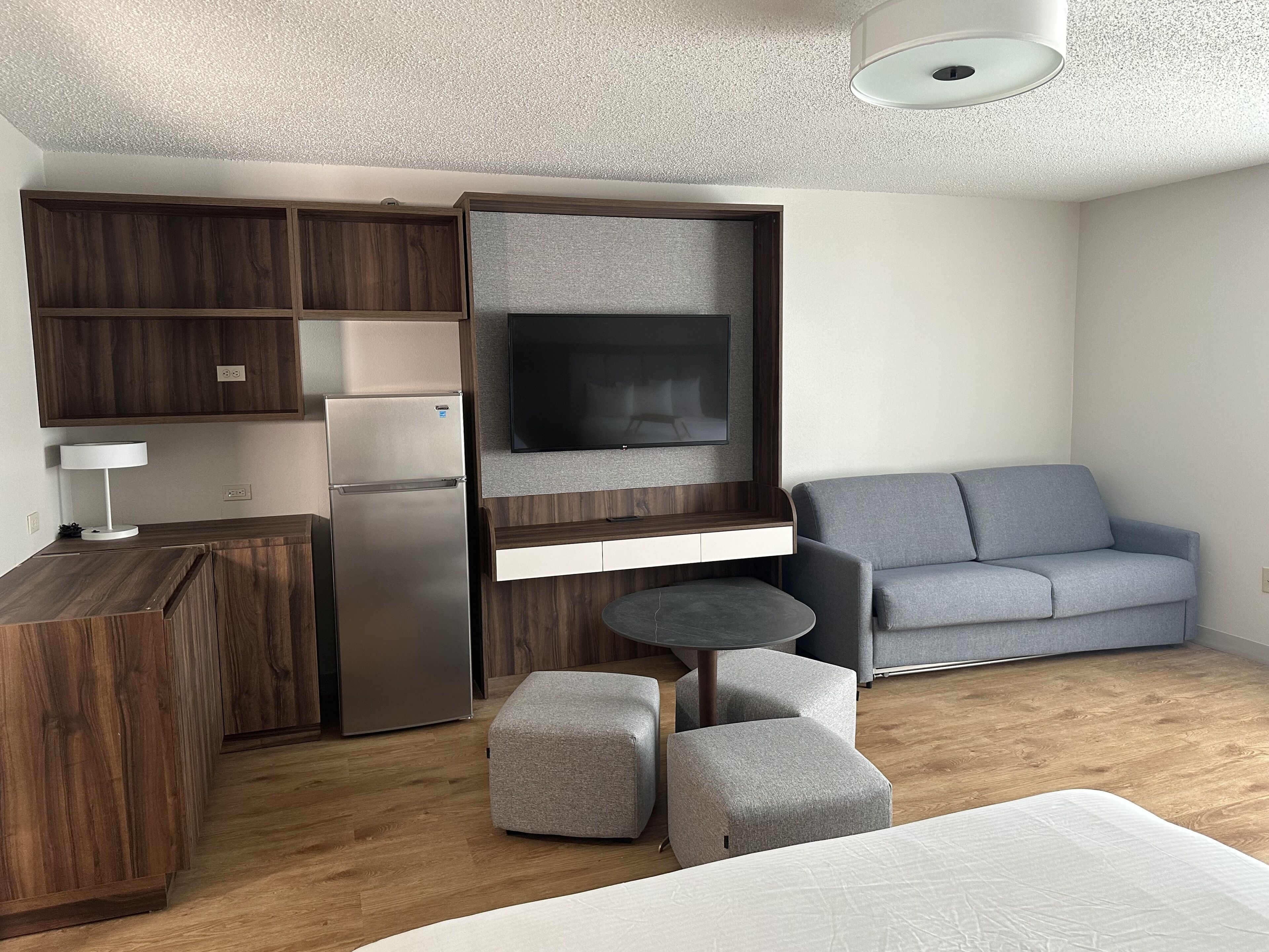 Un espacio habitable compacto con kitchenette, un televisor de pantalla plana, un sofá gris y una pequeña mesa redonda.