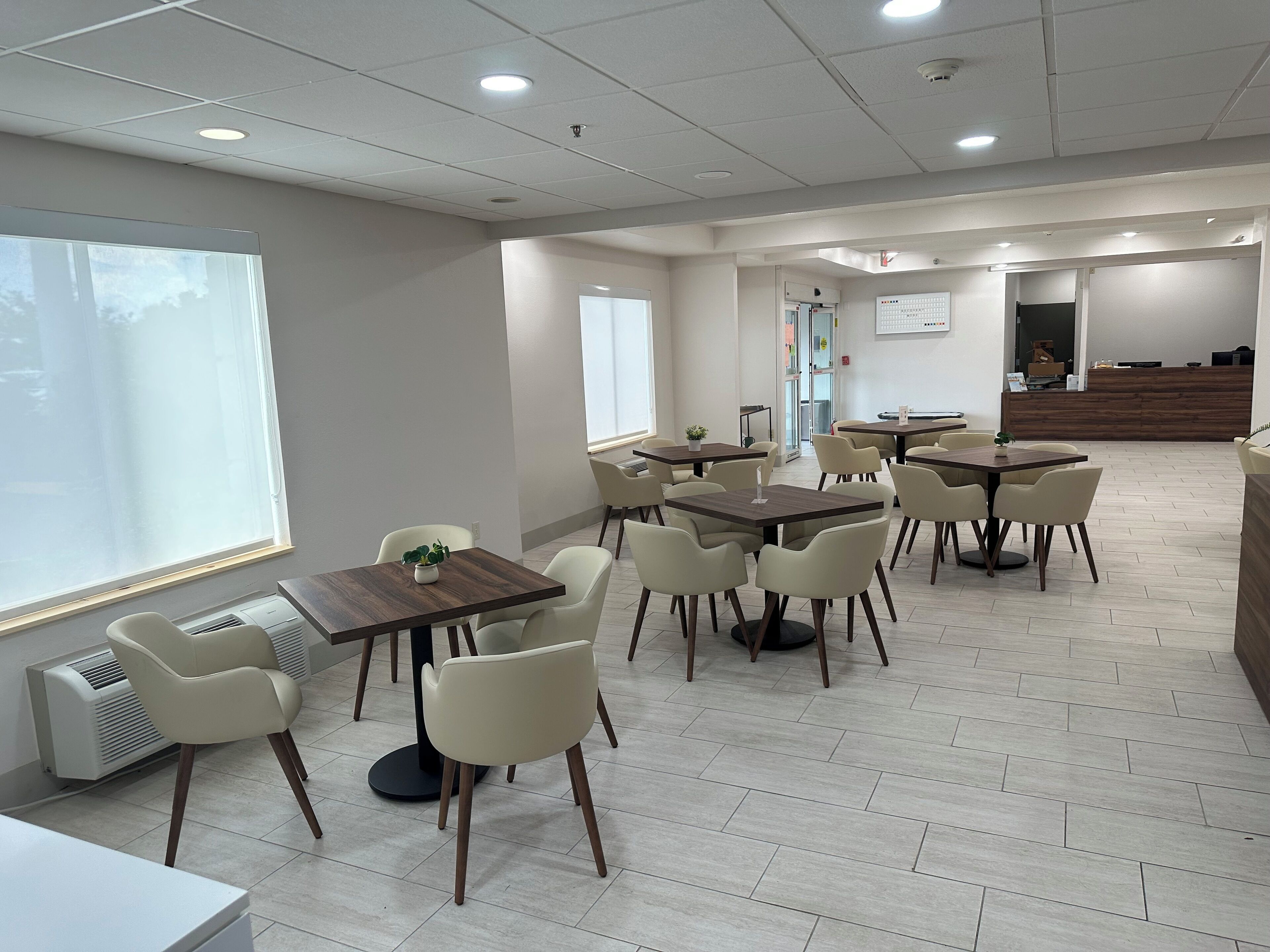 A modern dining area with multiple tables and chairs, a large window, and a reception desk in the background.