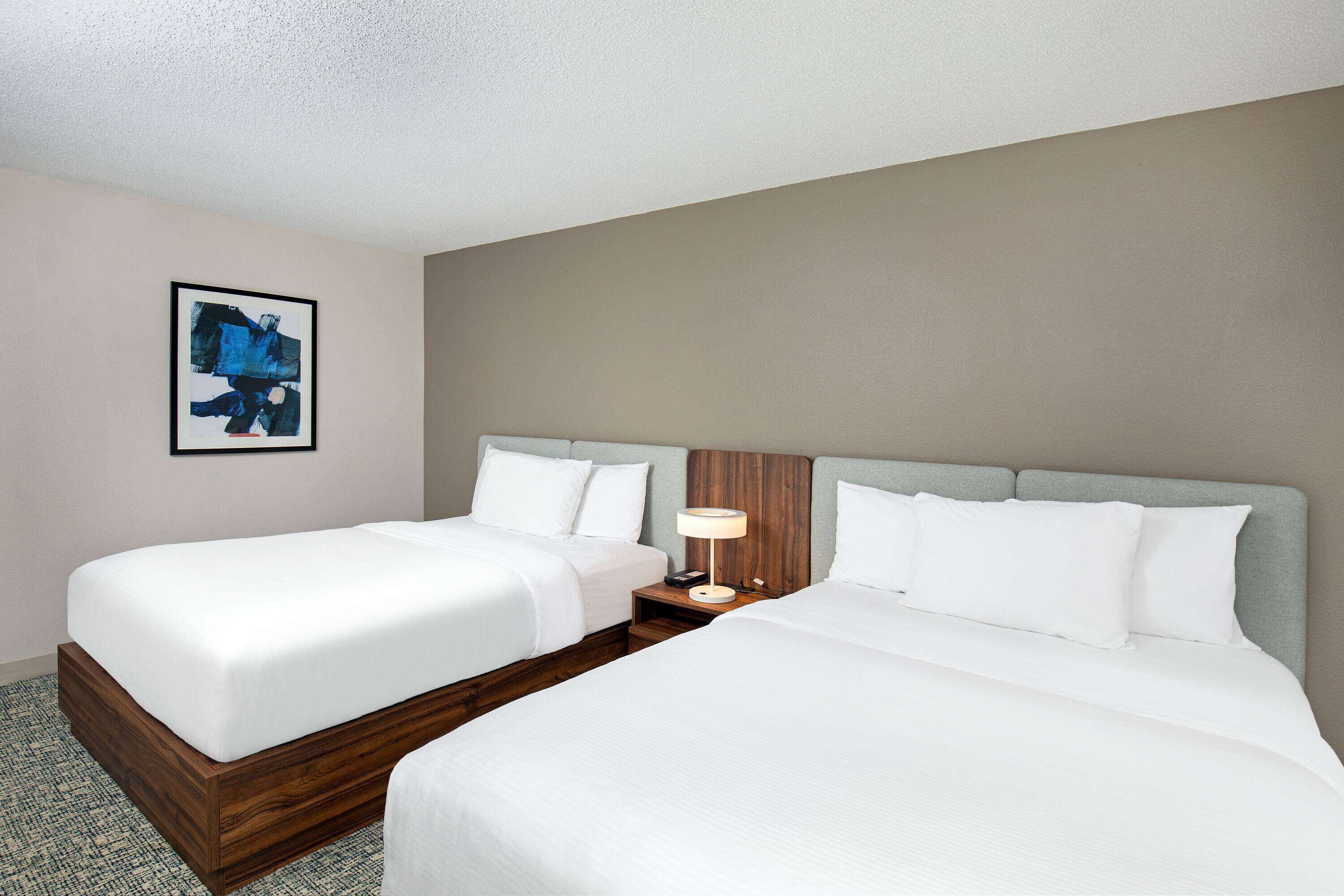 Habitación de hotel con dos camas, una mesita de noche con lámpara y un cuadro en la pared.