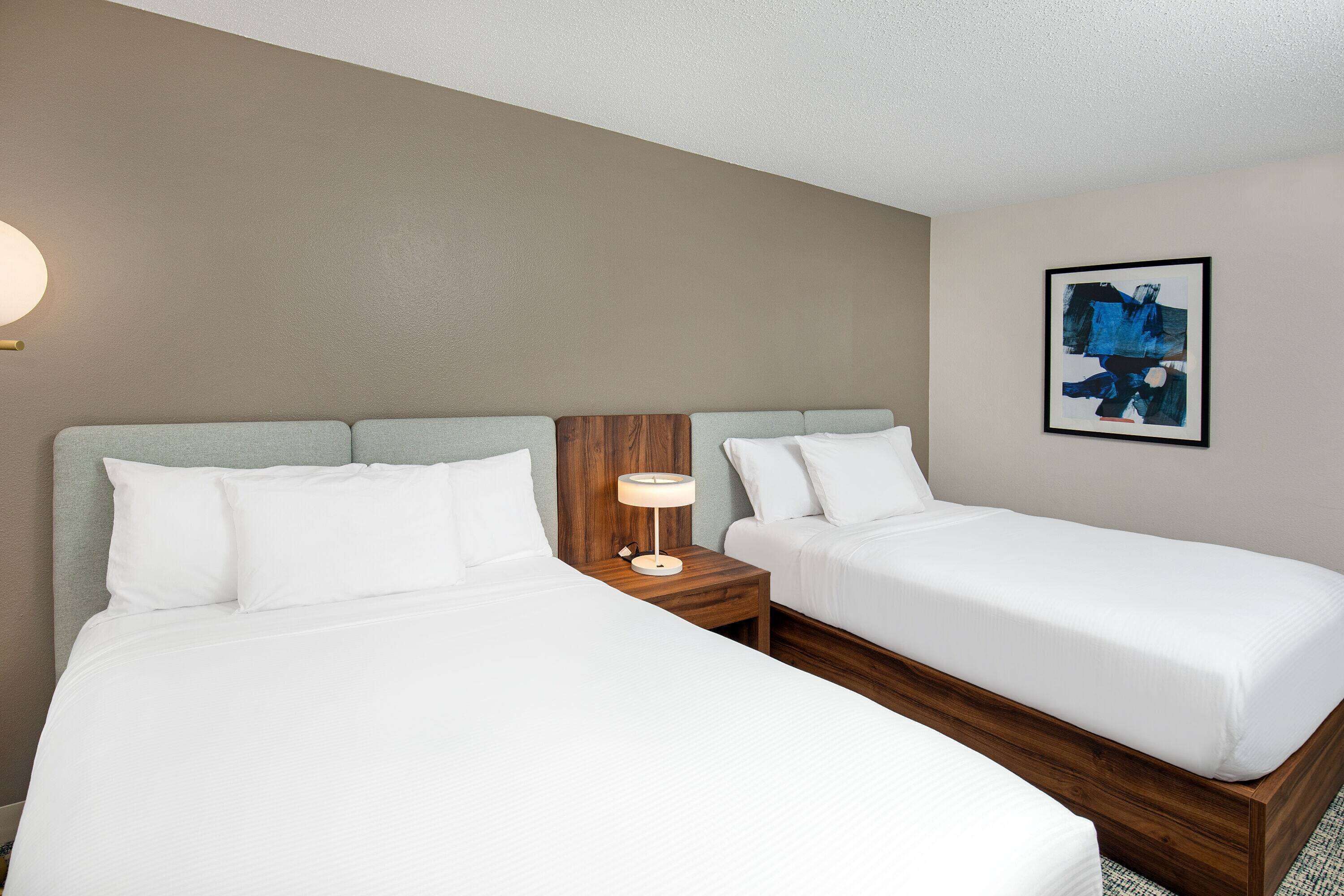 A hotel room with two single beds, a wooden headboard, a nightstand with a lamp, and a framed abstract painting on the wall.