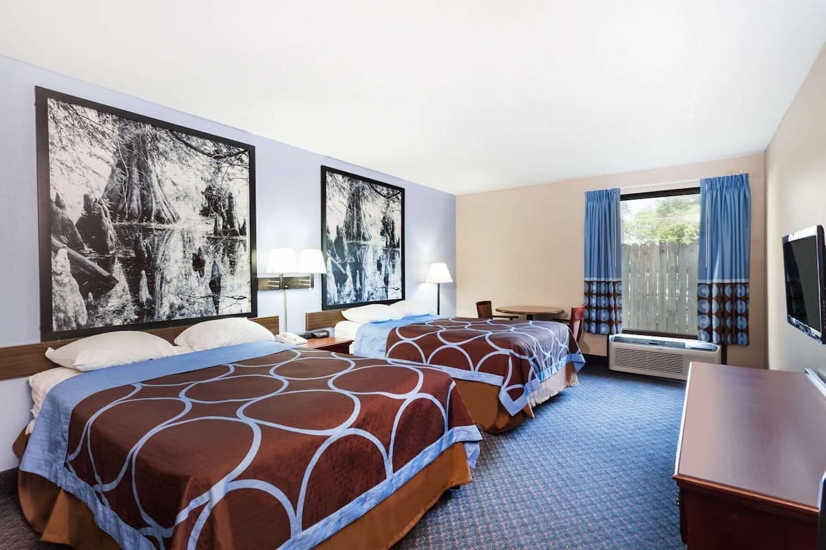 Una habitación de hotel con dos camas, un escritorio, un televisor y ventanas amplias con cortinas.