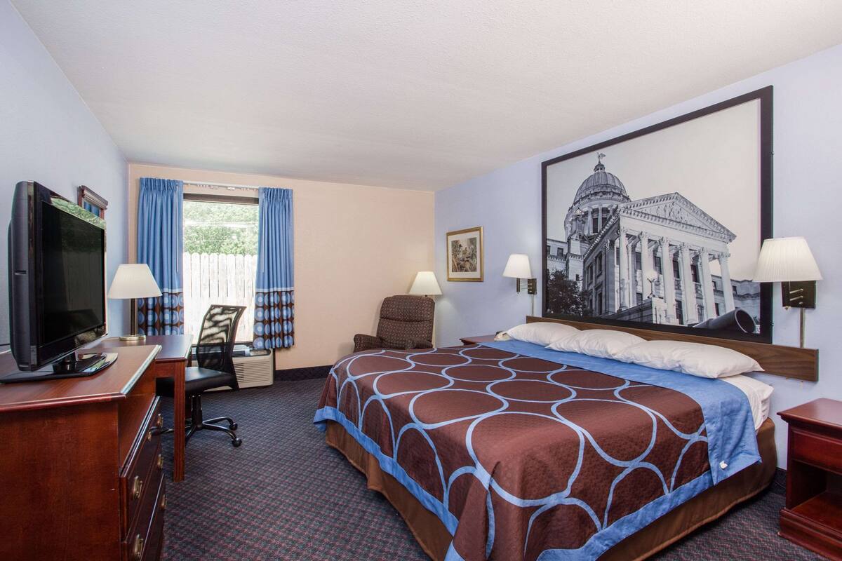 Habitación de hotel con una cama grande, un escritorio con televisor, dos sillas y un cuadro grande en la pared.