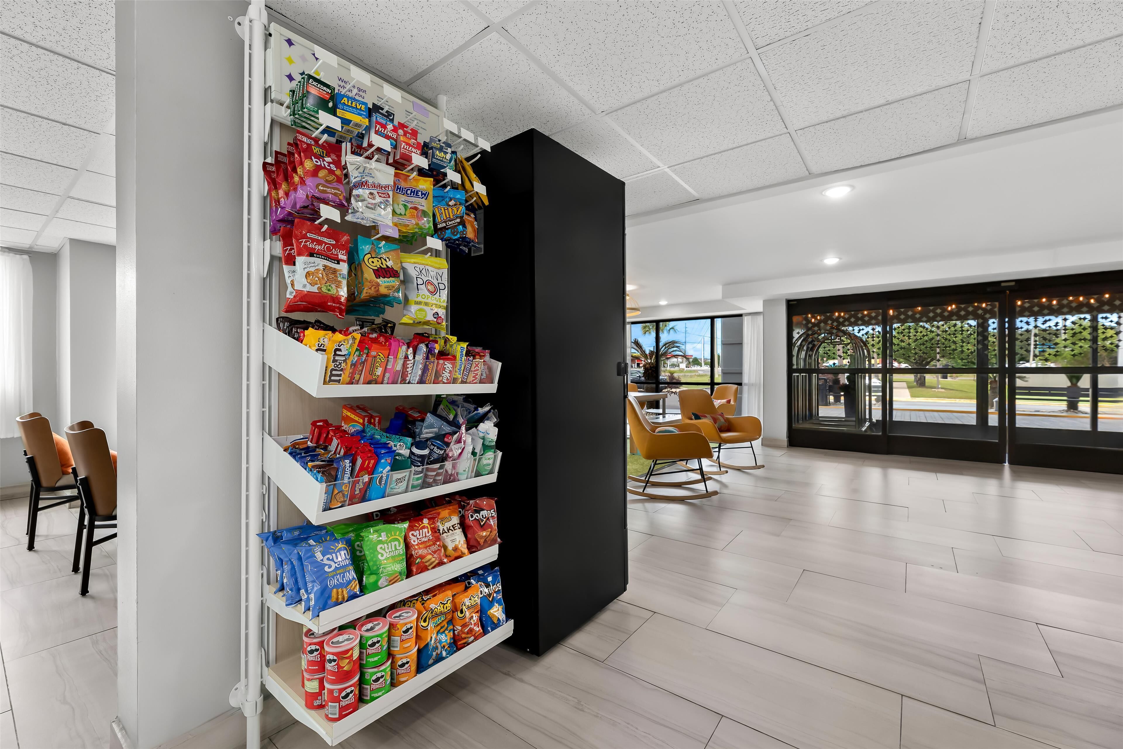 Ein modernes Interieur mit einem Snackautomaten, gefüllt mit verschiedenen Snacks und Getränken, einer Sitzecke mit Stühlen und großen Fenstern mit Blick ins Freie.