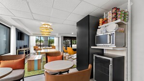 Una cocina moderna con microondas, refrigerador y estantes con alimentos. En la zona contigua hay mesas y sillas de comedor.