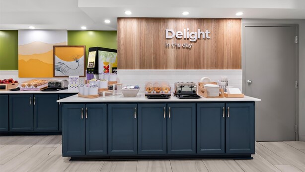 A modern kitchen area with a large island, a wooden accent wall with the text "Delight in the day," and various breakfast items displayed.