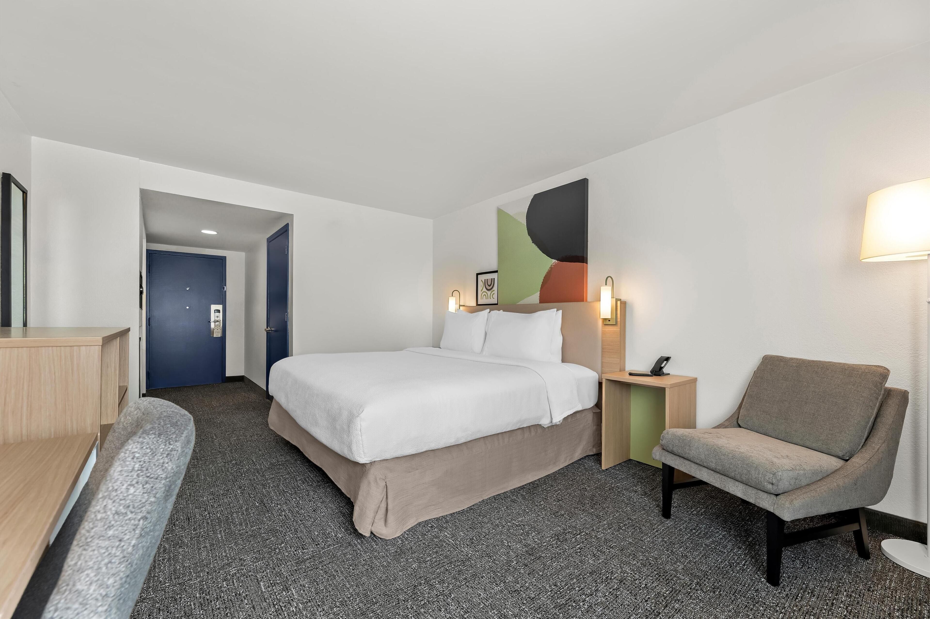 A hotel room with a bed, a desk, a chair, and a wall-mounted lamp.