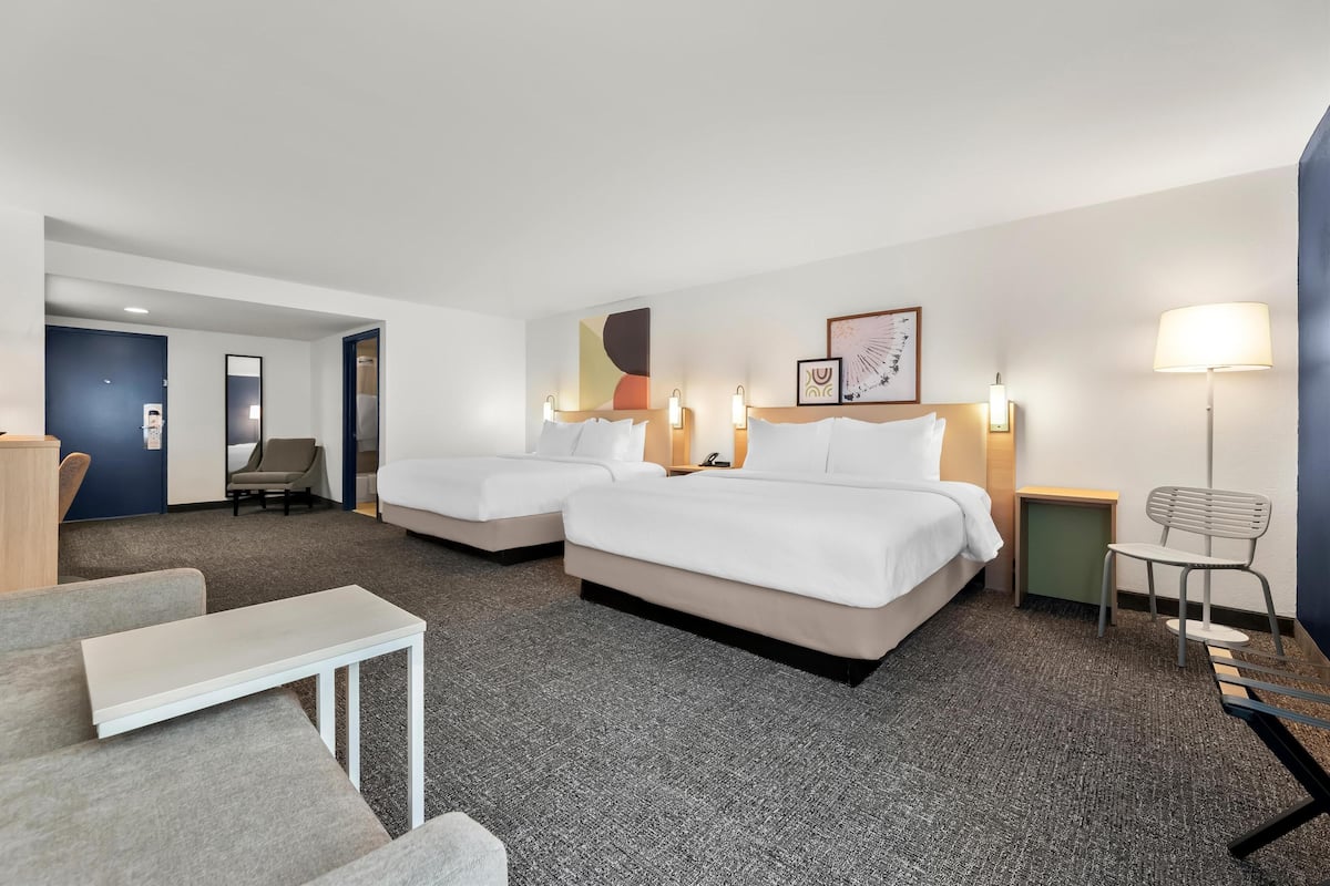 Hotelkamer met twee bedden, een kleine tafel, een stoel en een bureau. Er is een deur die naar een andere kamer leidt en een muur met kunstwerken.