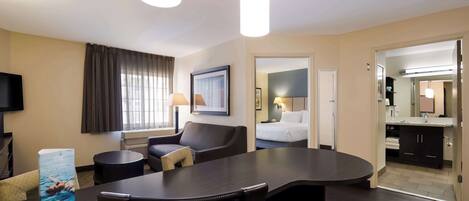 A hotel room with a dark wood table, a sofa, a TV, and a bed. There is a bathroom visible through an open door.