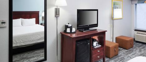 A hotel room with a bed, a desk with a television, a lamp, a mini-fridge, and a small seating area.