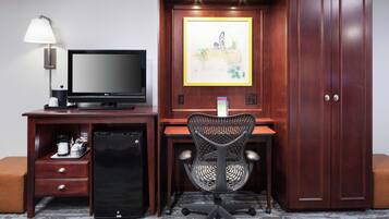 A hotel room with a wooden desk, a black office chair, a television, a refrigerator, a lamp, and a wardrobe.