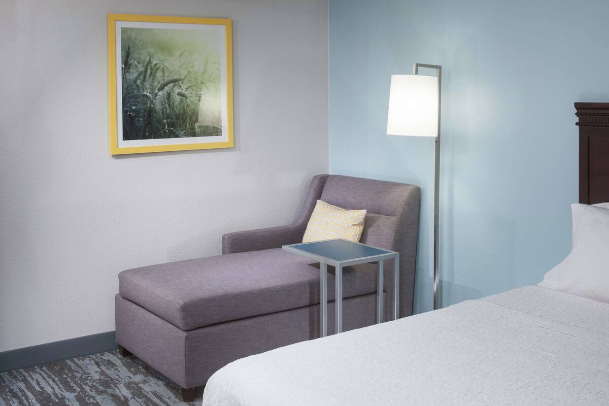 A hotel room with a bed, a small table, a chair, a lamp, and a framed picture on the wall.