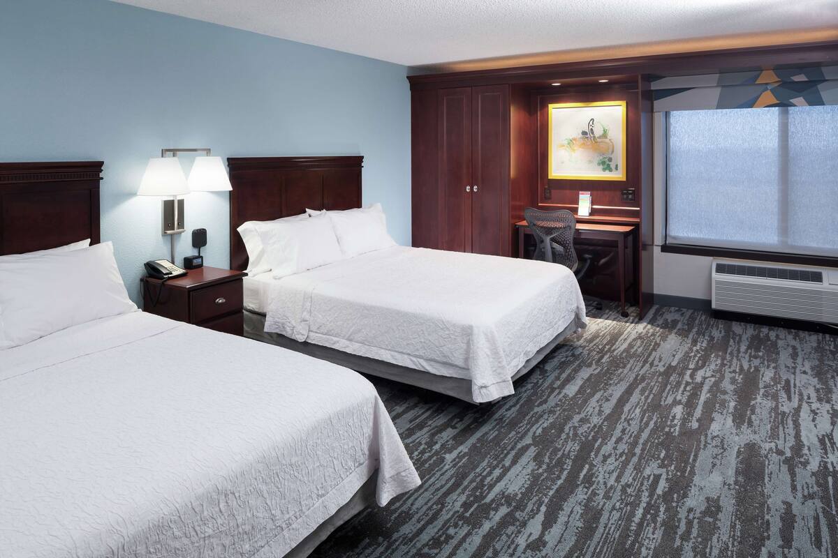 A hotel room with two beds, a desk, a chair, a lamp, a phone, and a painting on the wall.