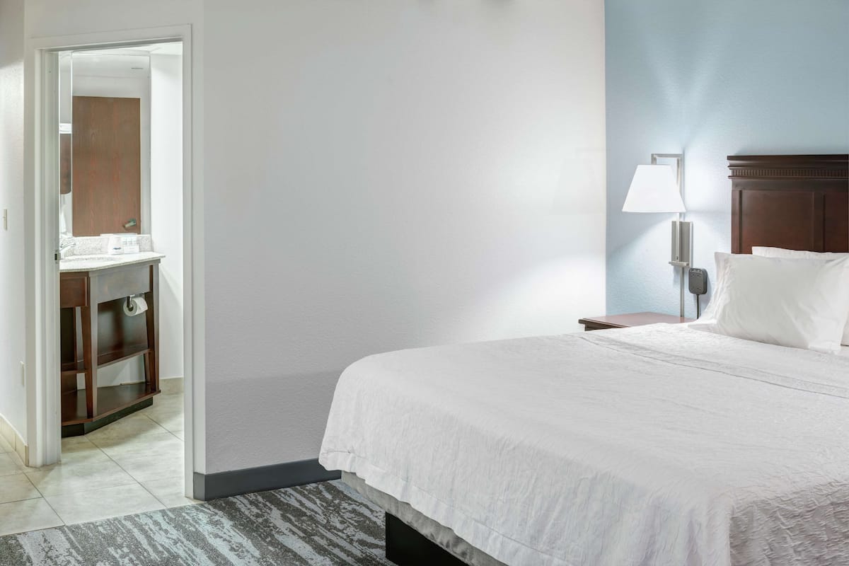 A hotel room with a bed, a nightstand, a lamp, a mirror, and a wooden cabinet.