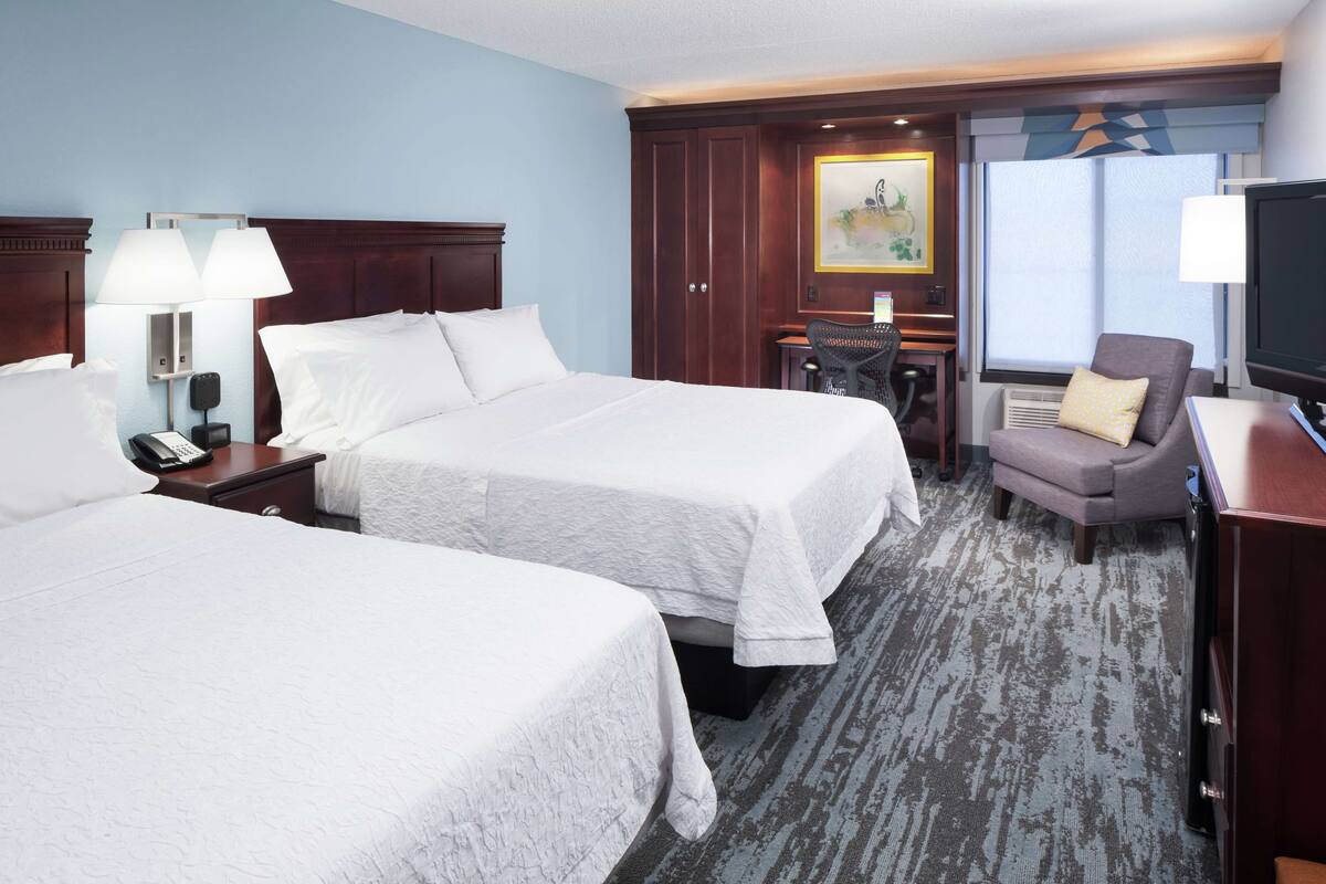 A hotel room with two beds, a desk, a chair, a TV, and a painting on the wall.