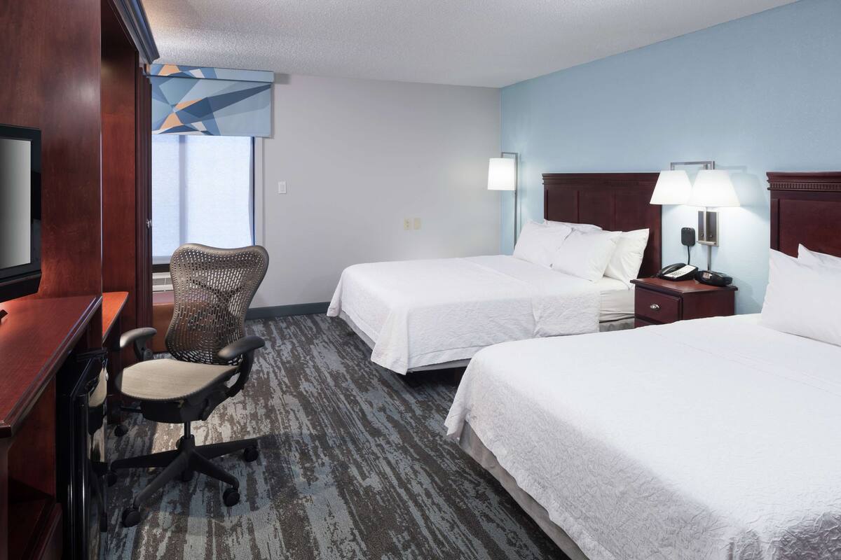 A hotel room with two beds, a desk with a chair, a television, and a lamp.