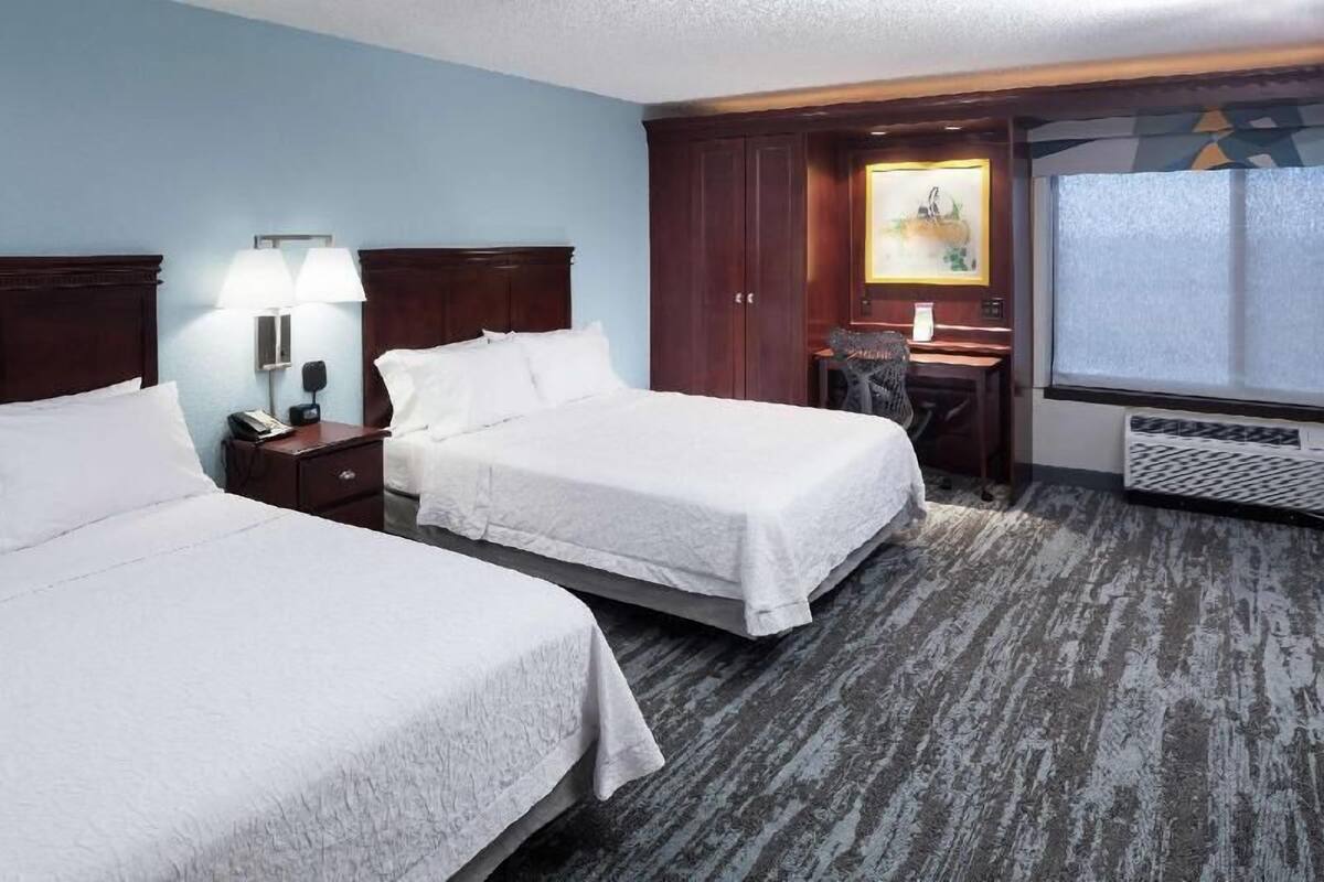 A hotel room with two beds, a desk, a chair, a lamp, and a painting.
