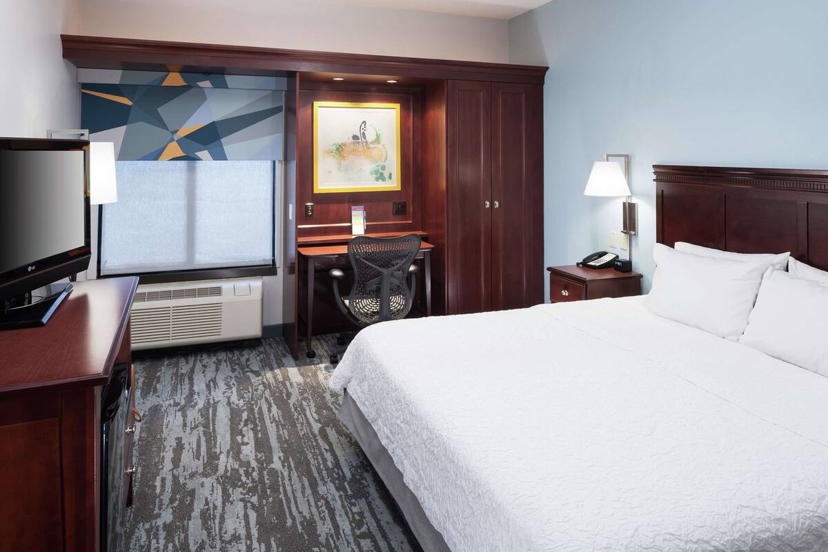 A hotel room with a large bed, a desk with a chair, a TV, a phone, and a painting on the wall.